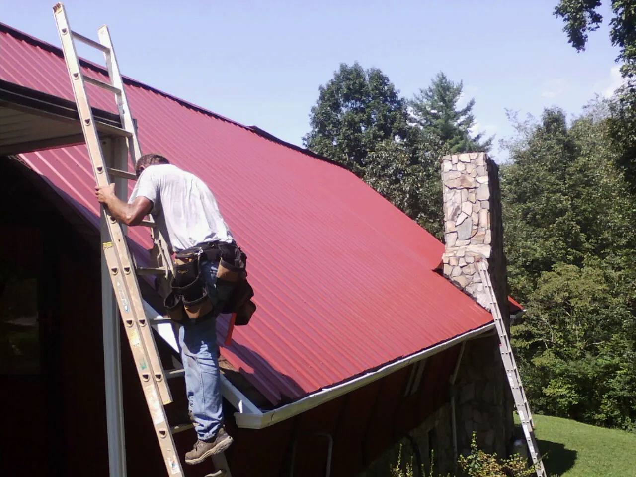 Roofer on a ladder beside a red metal roof and stone chimney — Soffit & Fascia Repair by Roofing Brothers in Brilliant